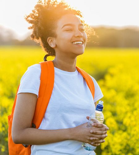 Outdoor portrait of beautiful happy laughing mixed race African American girl teenager female young woman with drinking water bottle in a field of yellow flowers at sunset in golden evening sunshine